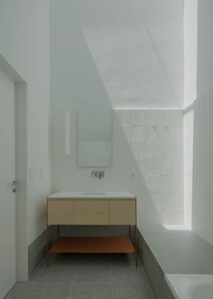 A minimalist bathroom with white ceramic tiles, concrete flooring, and a wood vanity.