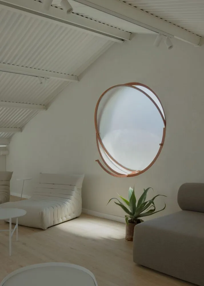 A modern living space featuring a white wall and light-colored wood flooring, with a round window and potted plant.