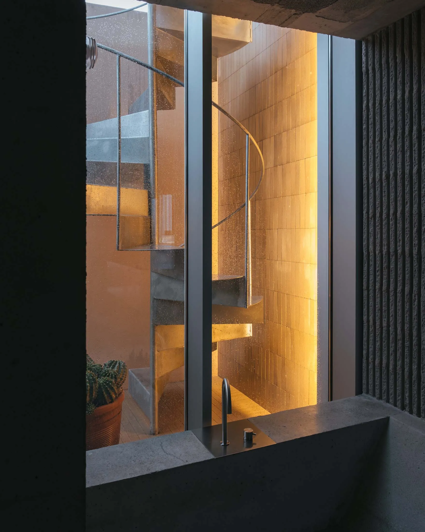 A modern, spiral staircase with metal railings illuminated by warm lighting, seen through a window with condensation.