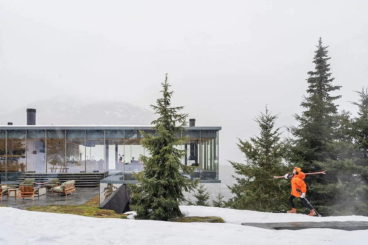 現代住宅建築採用玻璃牆面與鋼材點綴,周圍環繞雪景與常青樹,一位滑雪者正滑過
