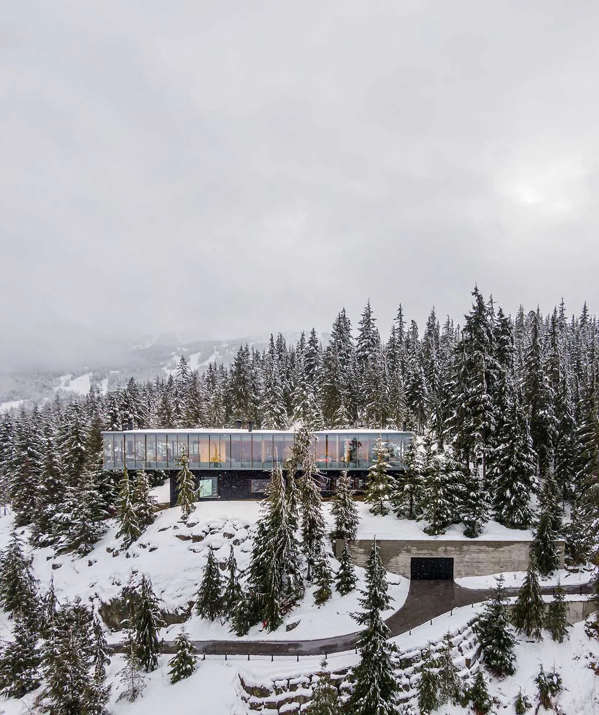 現代玻璃小屋坐落於雪覆蓋的山坡上，周圍環繞著常青樹。
