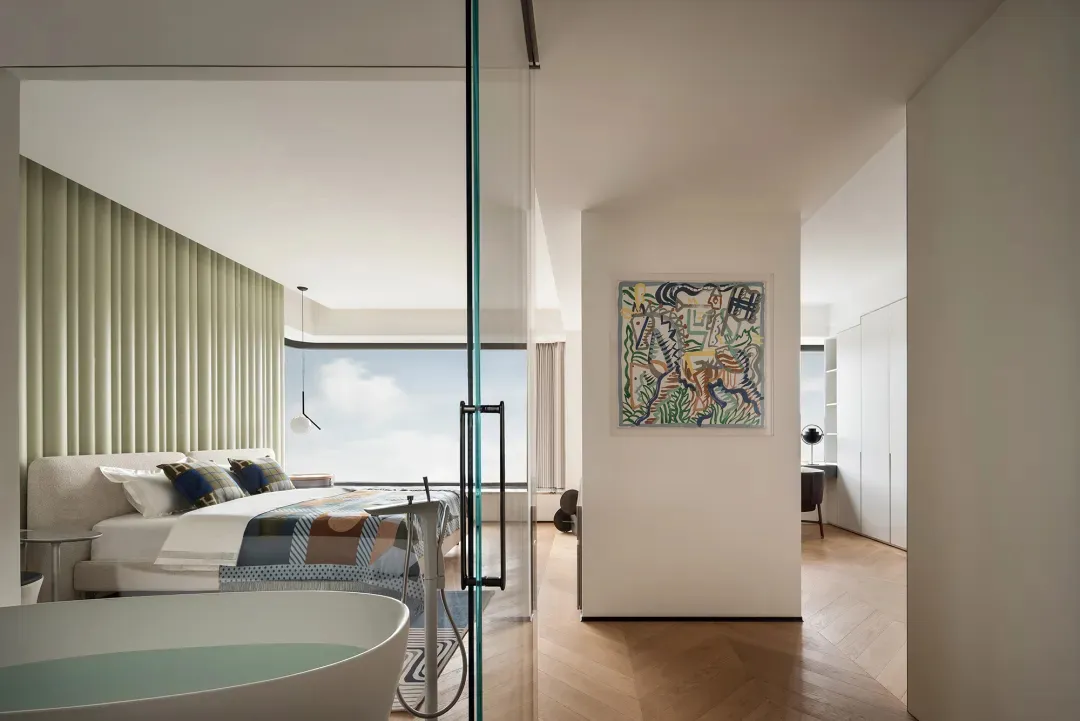 A minimalist bedroom with a large window, a queen-sized wooden bed with a patterned bedspread, and a glass bathtub.