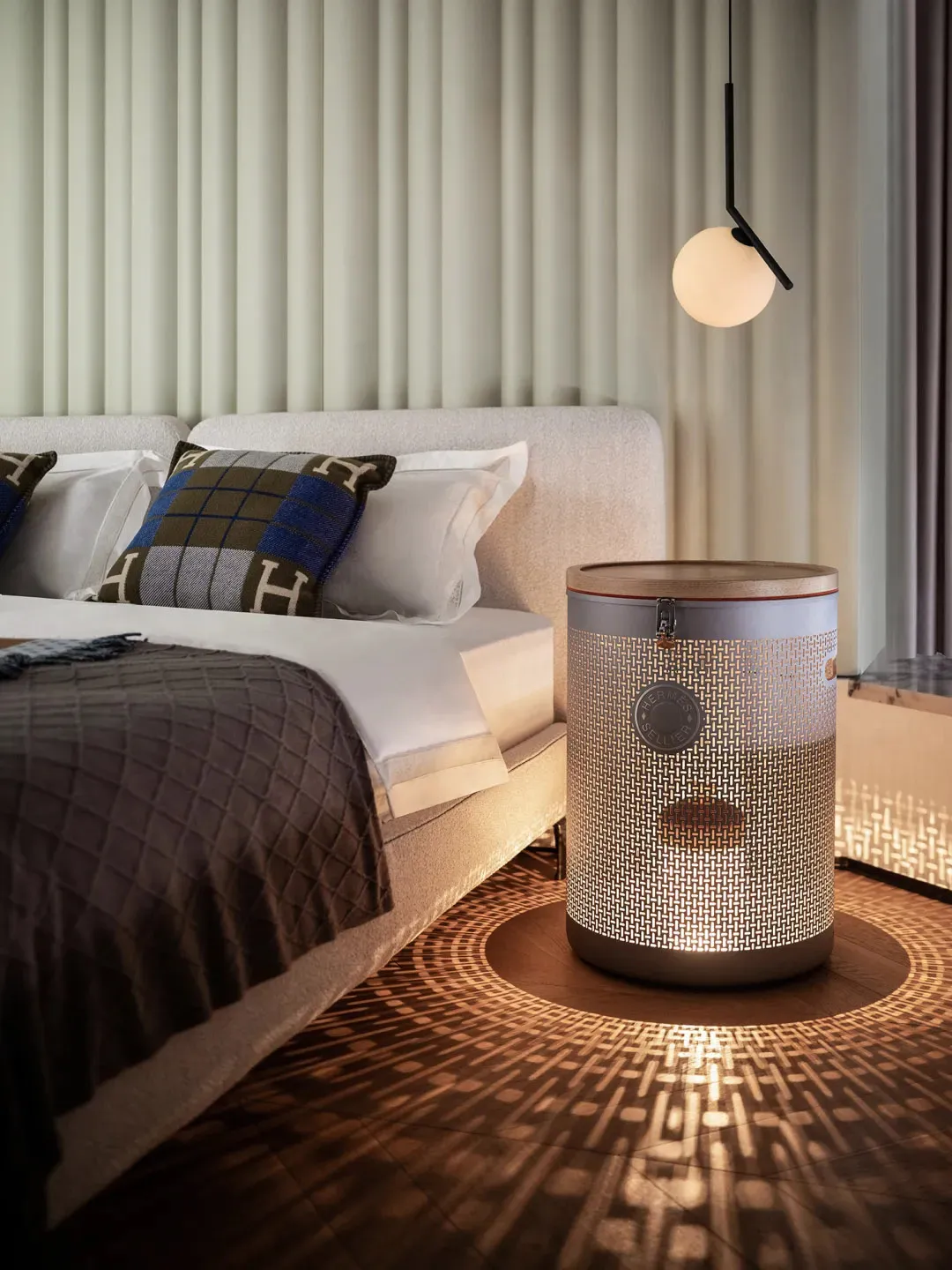 A modern minimalist bedroom with a bed, patterned textiles, and a lit cylindrical nightstand.