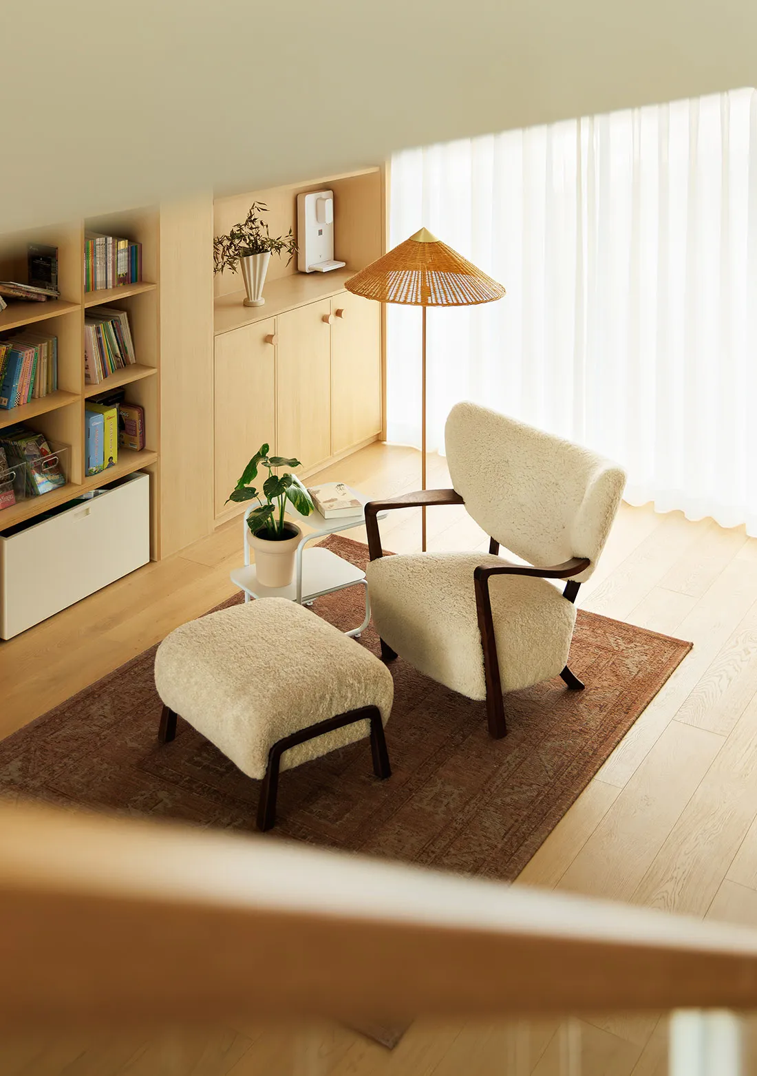 A cozy living room with a comfortable armchair and matching ottoman, wooden flooring, and a side table with a plant and books