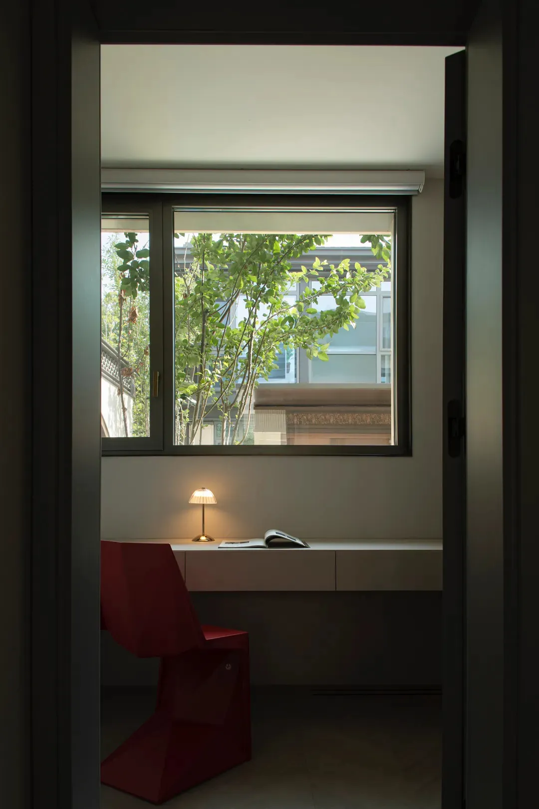 A modern workspace with a metal desk and a red chair, featuring a lamp and greenery visible through a window.