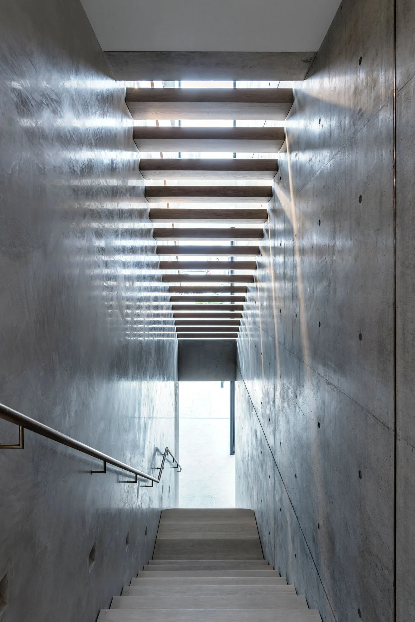 A modern staircase with wooden steps and concrete walls, illuminated by natural light from above.