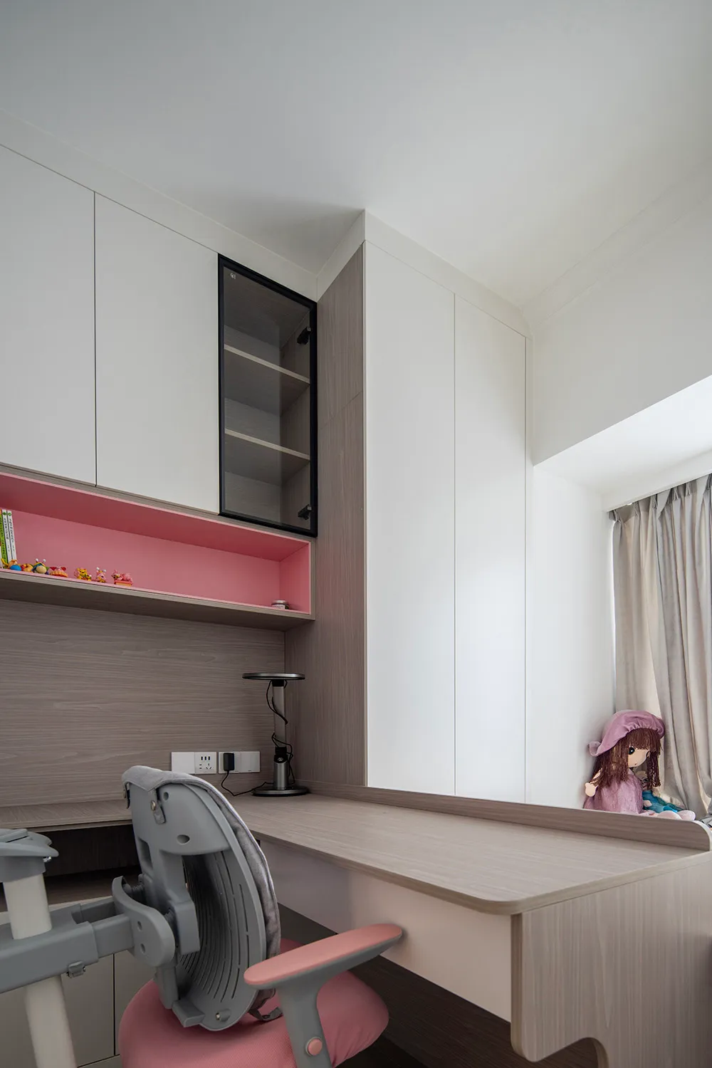 A modern study room with a wooden desk, grey chair, and white and pink cabinets.