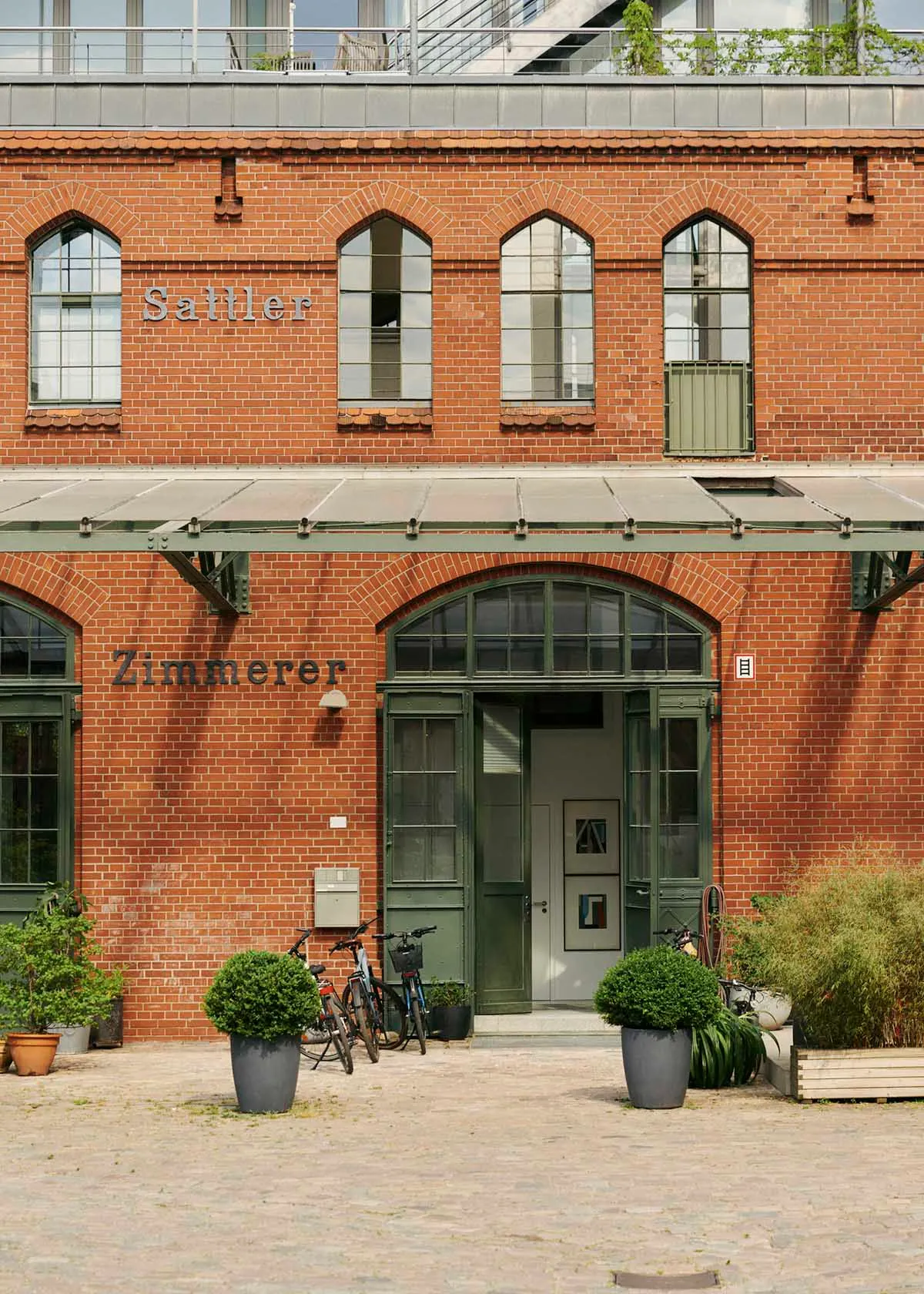 A rustic, industrial building with red brick walls and green window frames, featuring the words 'Sattler Zimmerer' above the
