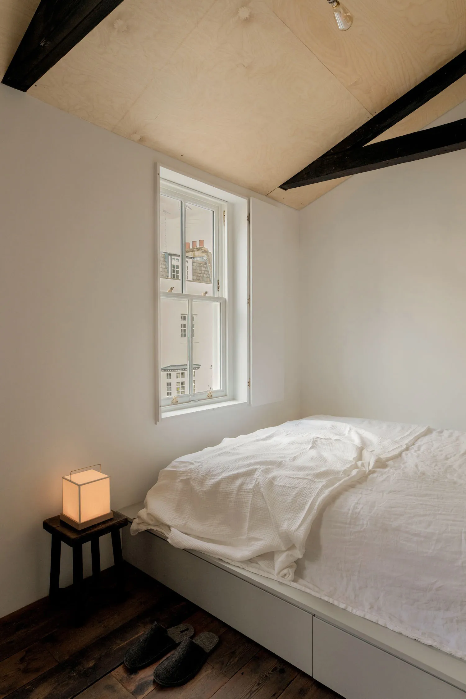 A minimalist bedroom with a white bed, wooden floor, and natural light coming through the window.