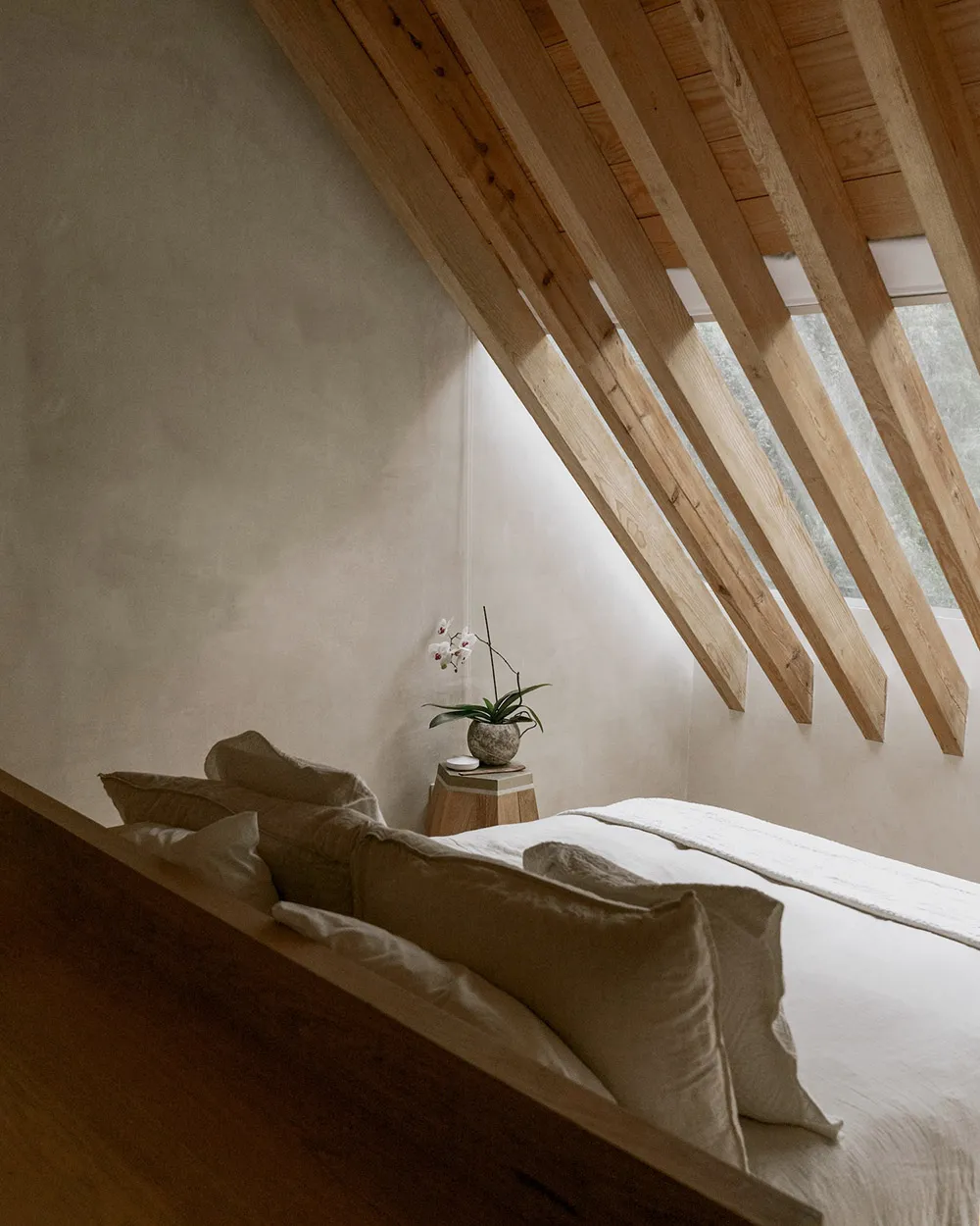 A minimalist bedroom with a wooden roof and white bedding, featuring a small white orchid on a wooden stool.
