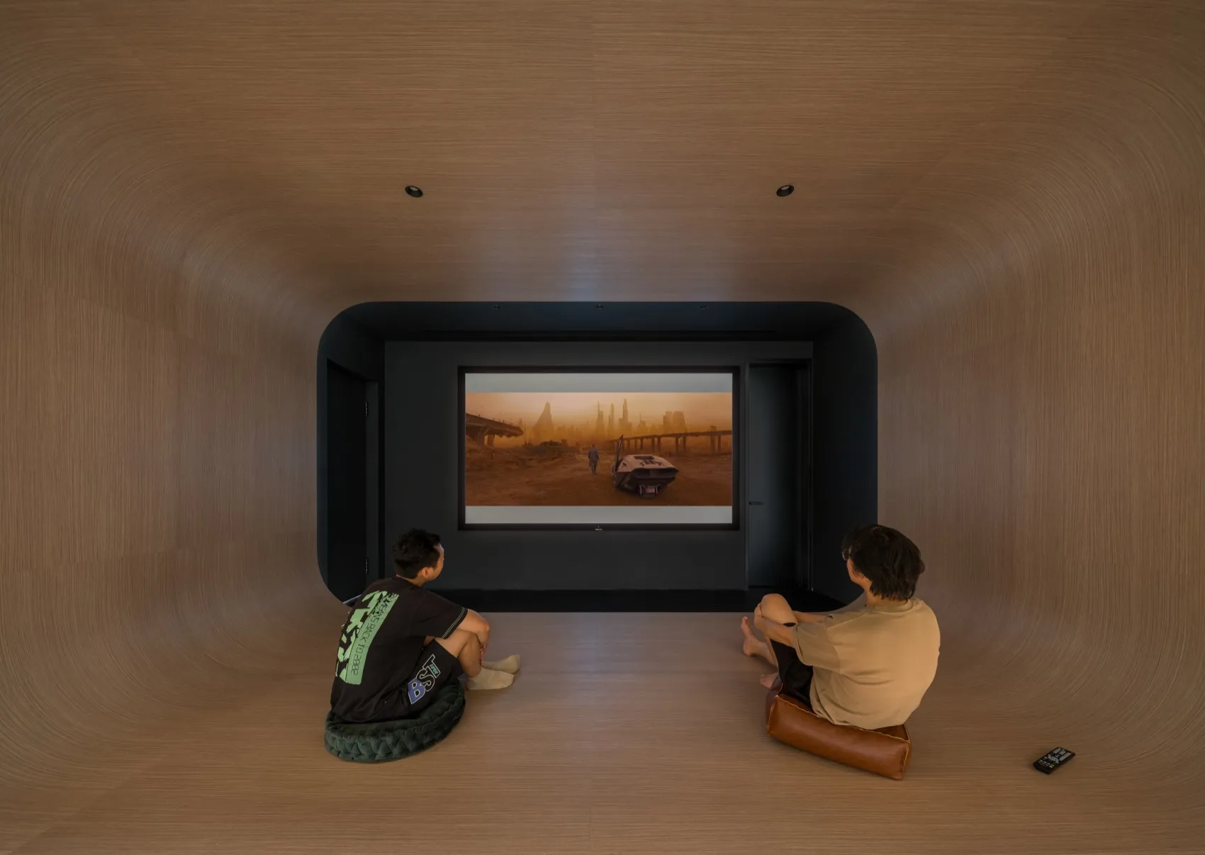 A minimalist home theater setup with two people sitting on floor cushions and watching a movie on a large screen.