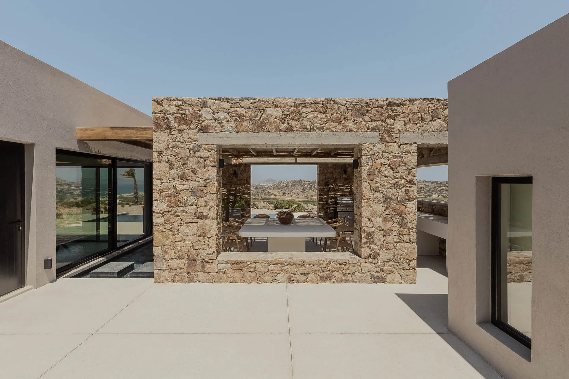 Modern minimalist patio with stone walls and concrete floor, featuring a dining table and chairs under an open-air roof, over