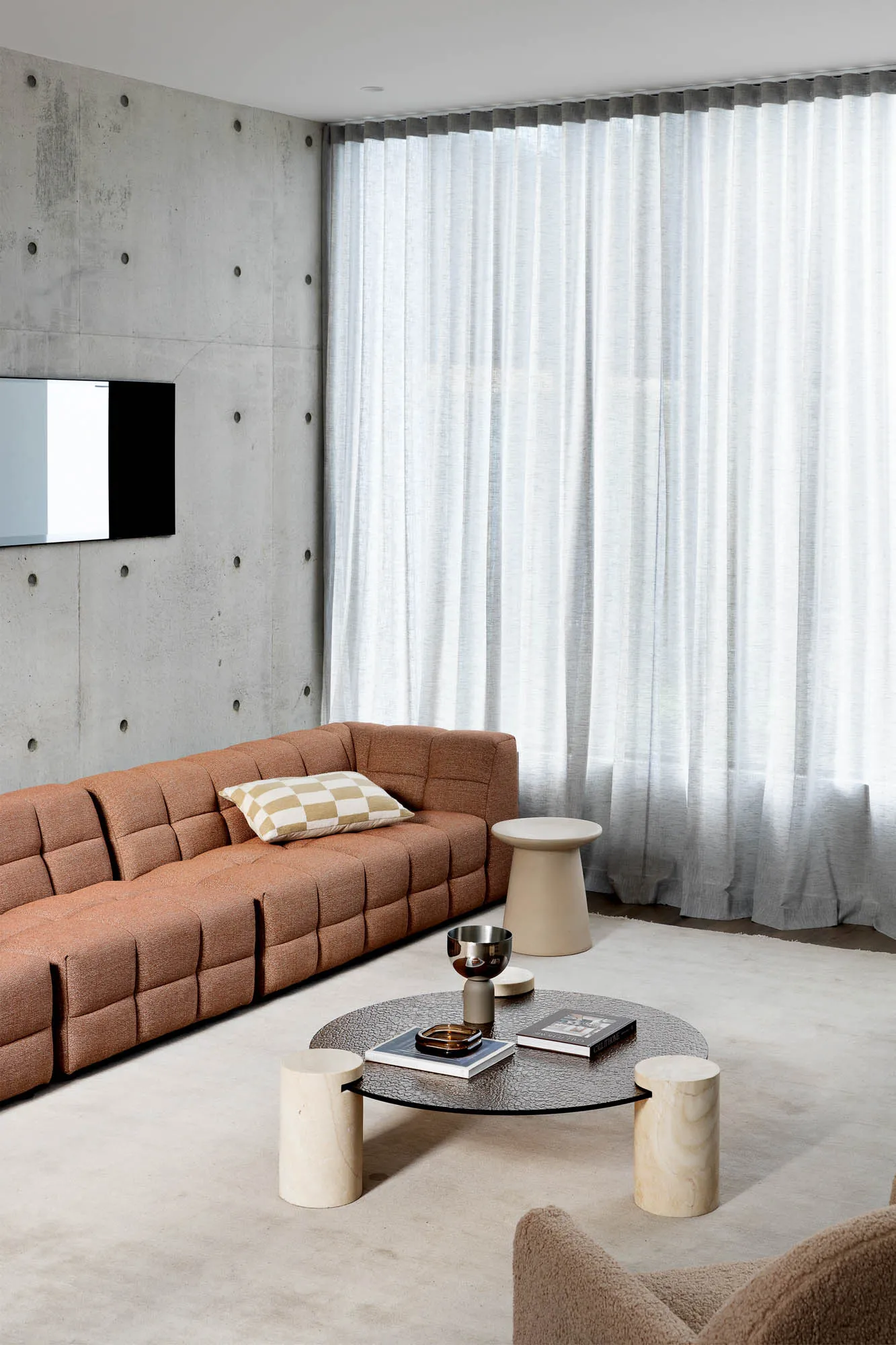 A modern living room with a brown modular sofa, cream-colored marble coffee table, and large windows with sheer curtains.