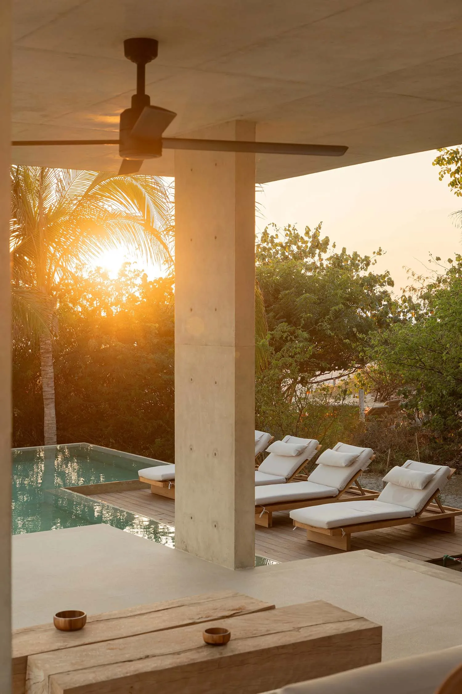 Modern outdoor lounge area with concrete structure, pool, lounge chairs, and sunset view through palm trees.