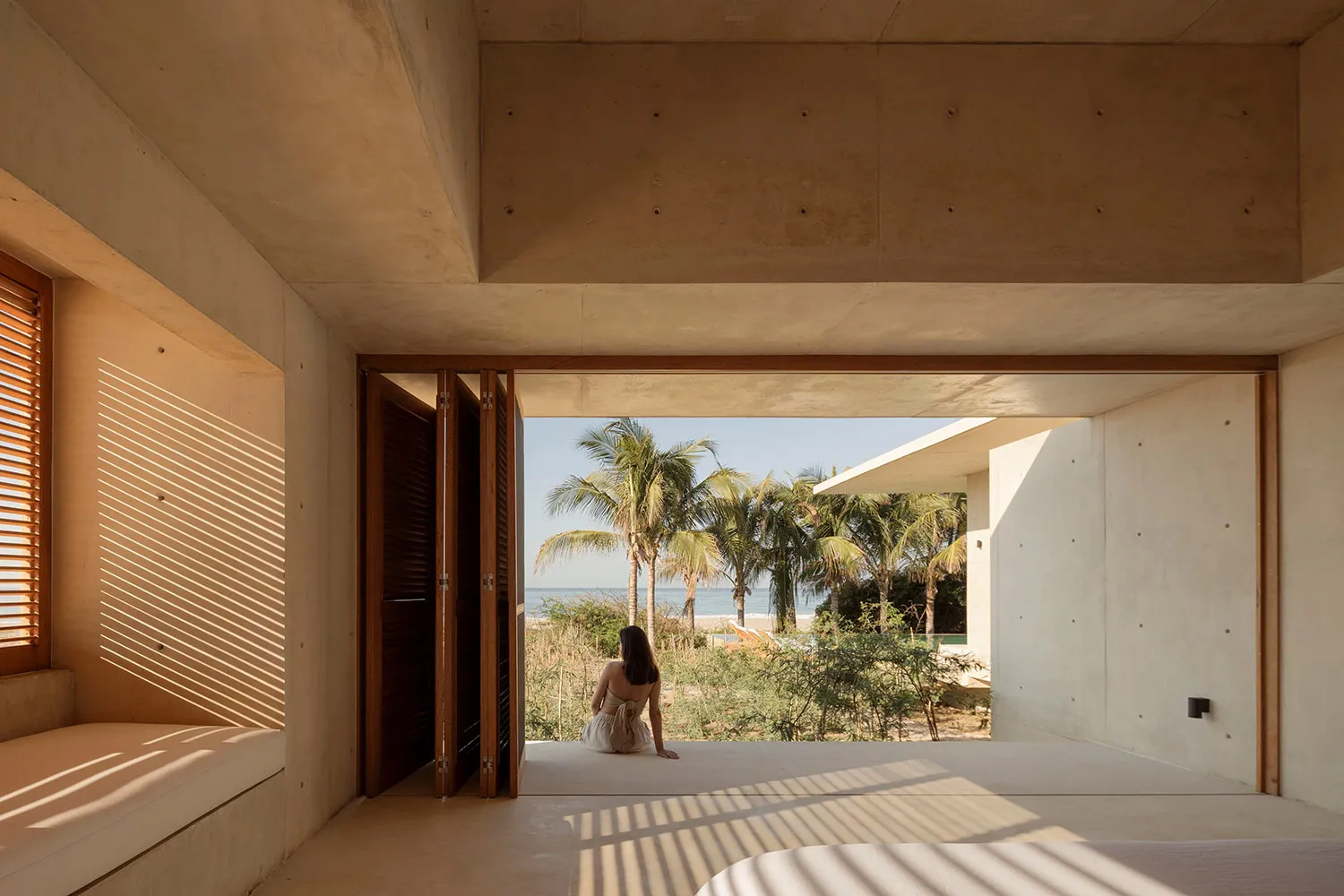 A modern residential bedroom with concrete walls and floor, featuring a view of the beach through an open window.