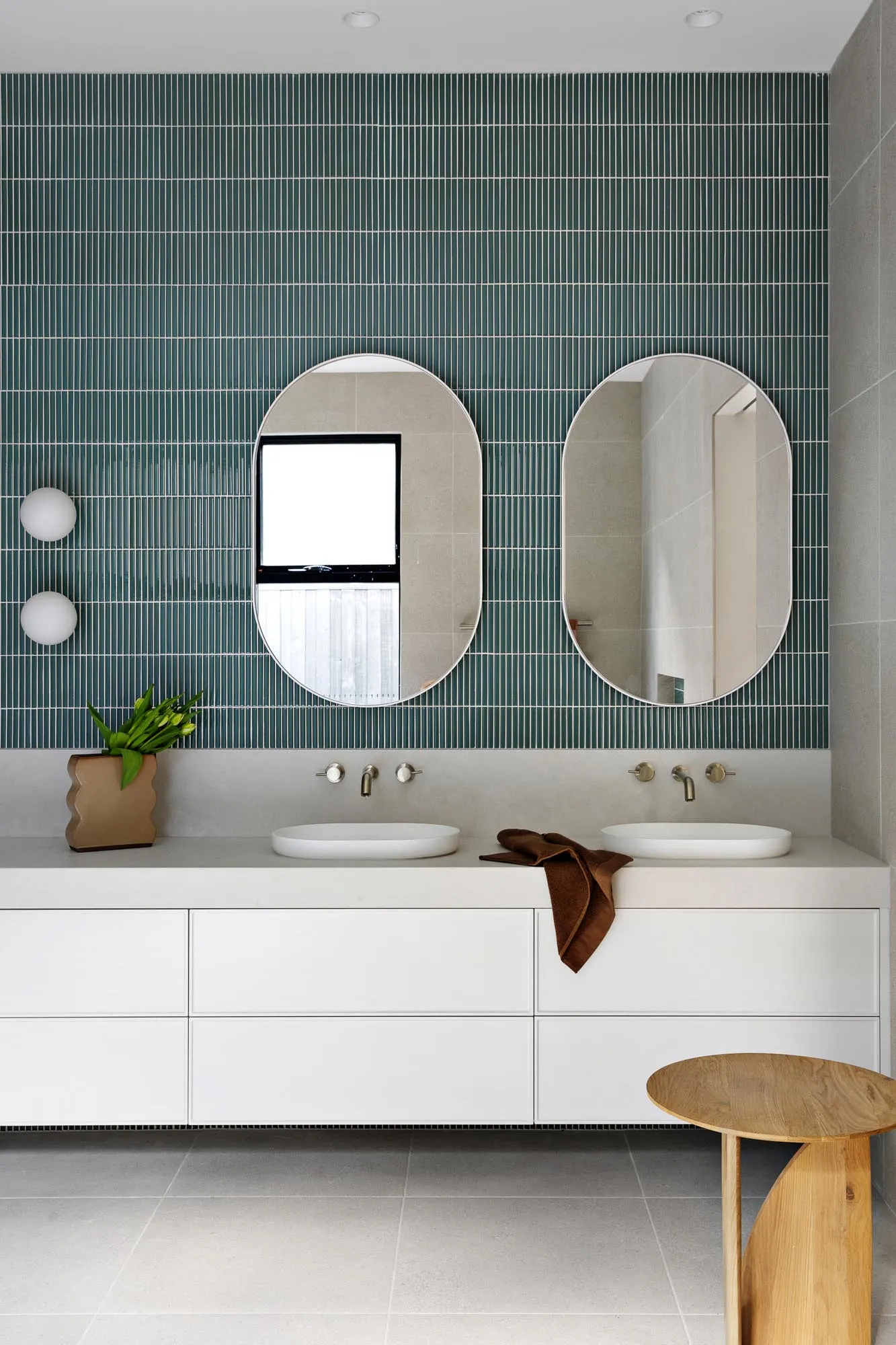 Modern bathroom with green mosaic tile wall, white concrete floating vanity, brass faucets, and a wooden stool.