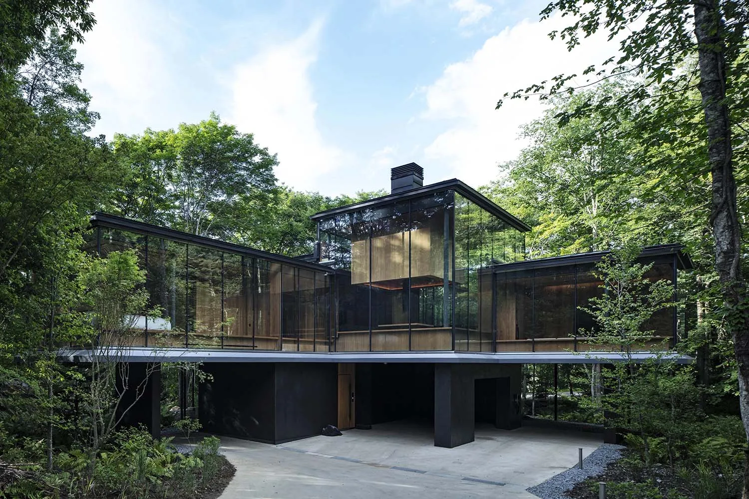 A modern, glass-fronted residential house surrounded by lush green trees and foliage.