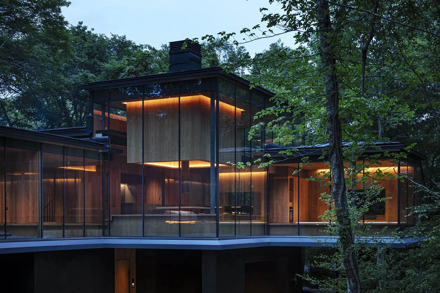 A modern glass house surrounded by trees with warm indoor lights visible through the glass walls.