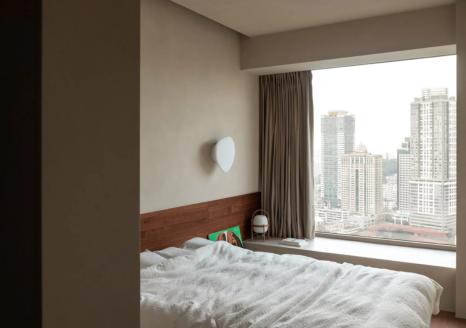 A minimalist bedroom with a large window overlooking the city, featuring a blank white blanket on the bed and a wall sconce.