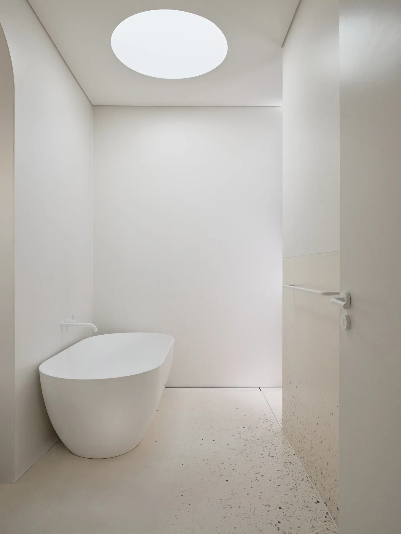 A minimalist bathroom with a white, oval-shaped bathtub, a terrazzo floor, and a circular skylight.