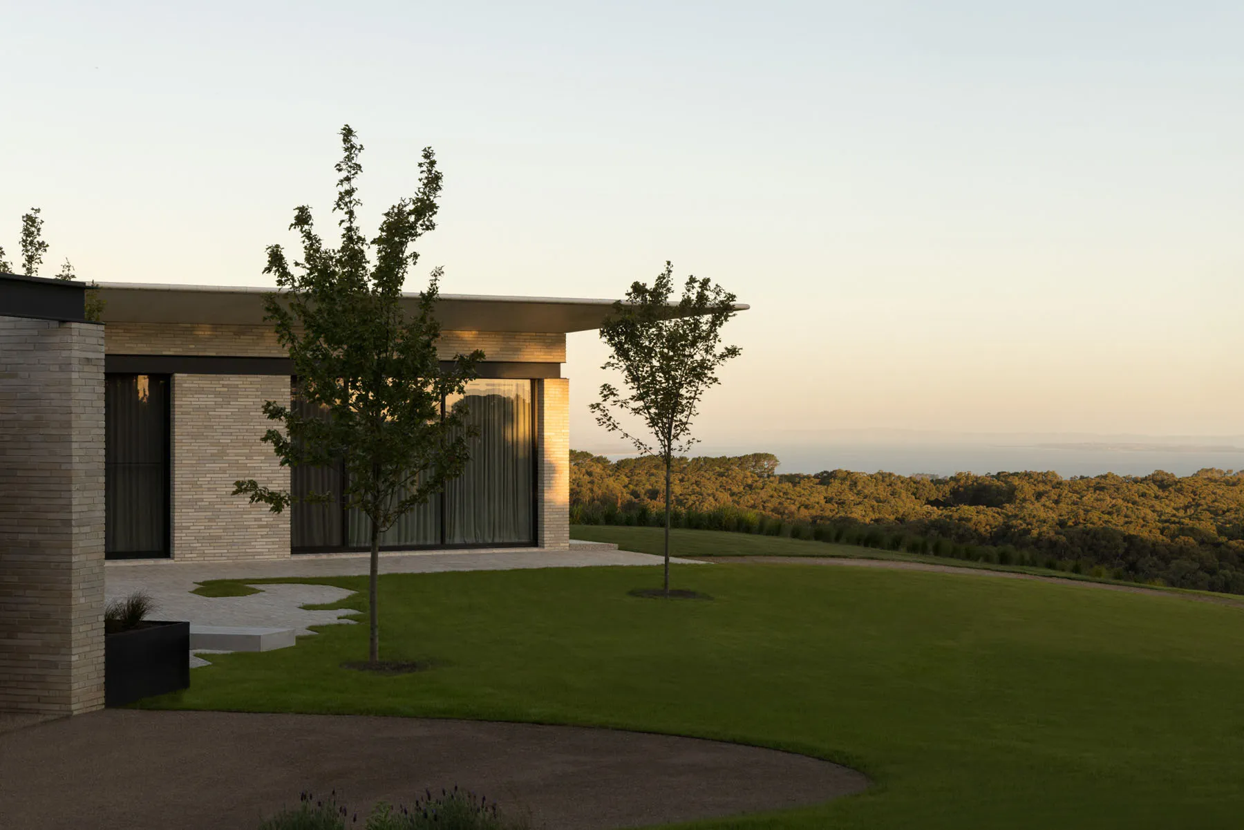 Modern residential exterior with brick walls, glass windows, and a landscaped yard overlooking a forested area during sunset.