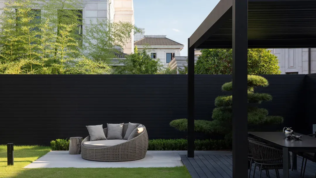 A minimalist outdoor space with black walls, featuring a round wicker daybed, a dining table with chairs, and greenery.