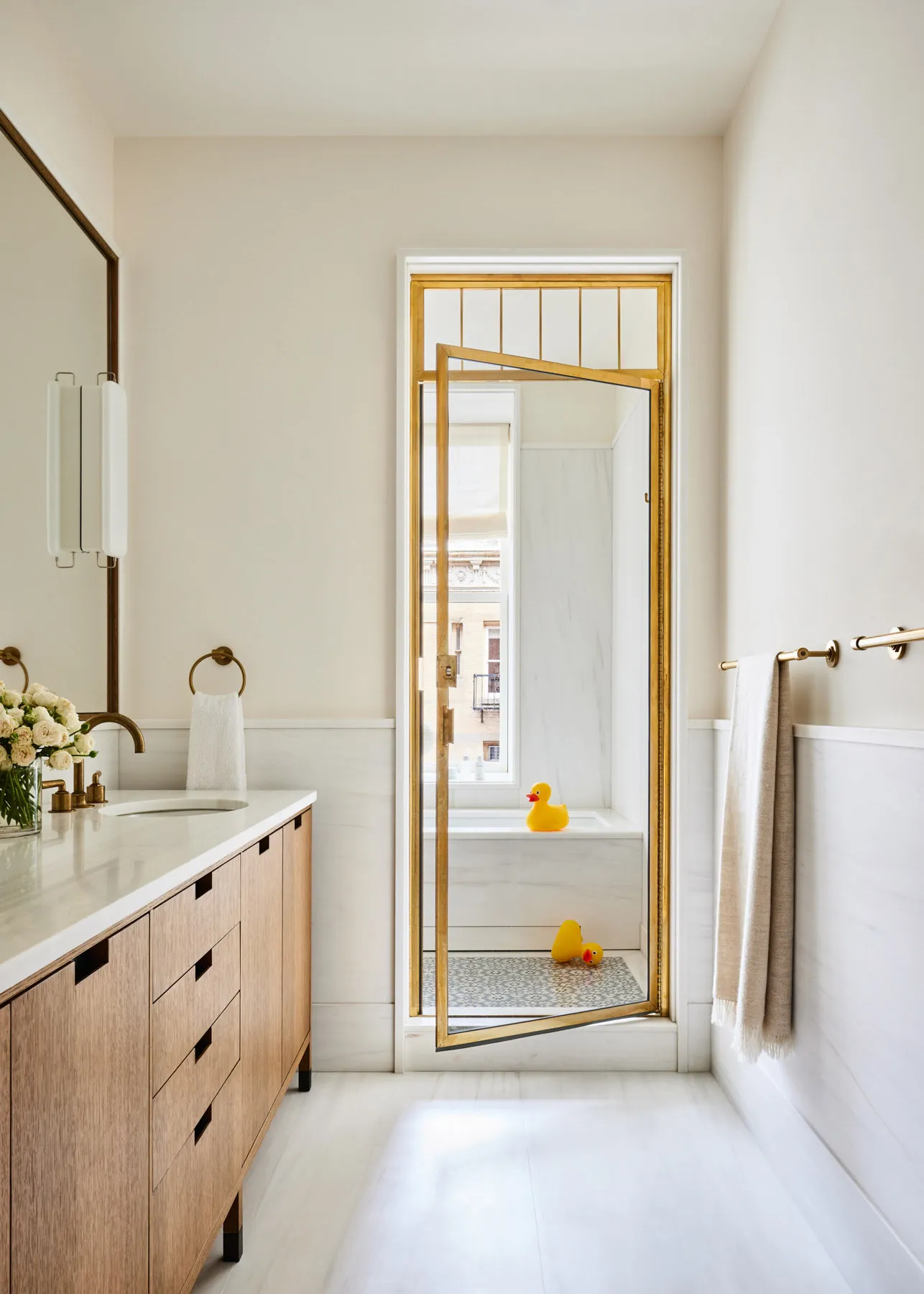 A minimalist white bathroom featuring a wooden vanity, marble countertops, and gold accents.