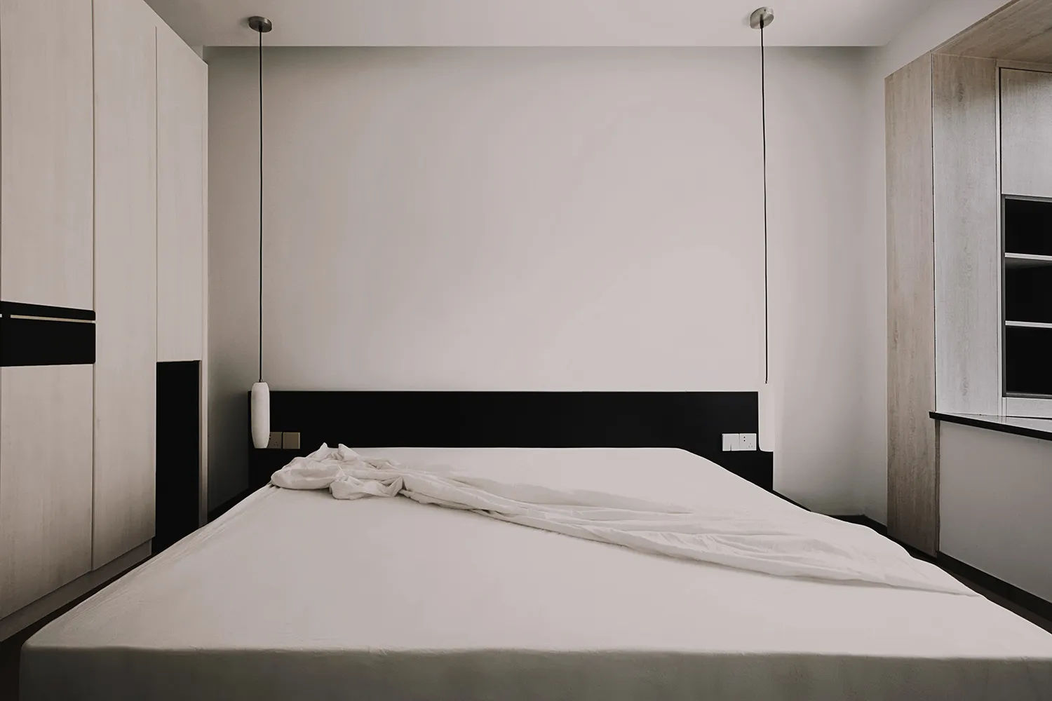A minimalist bedroom with a white textured bedspread and black headboard against a plain wall with hanging lamps.