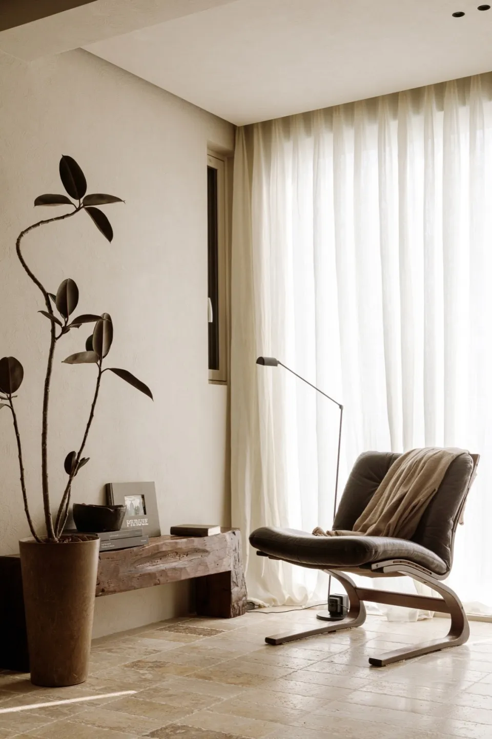 A modern living room with a black leather armchair, white curtains, and a potted plant in a tall vase.