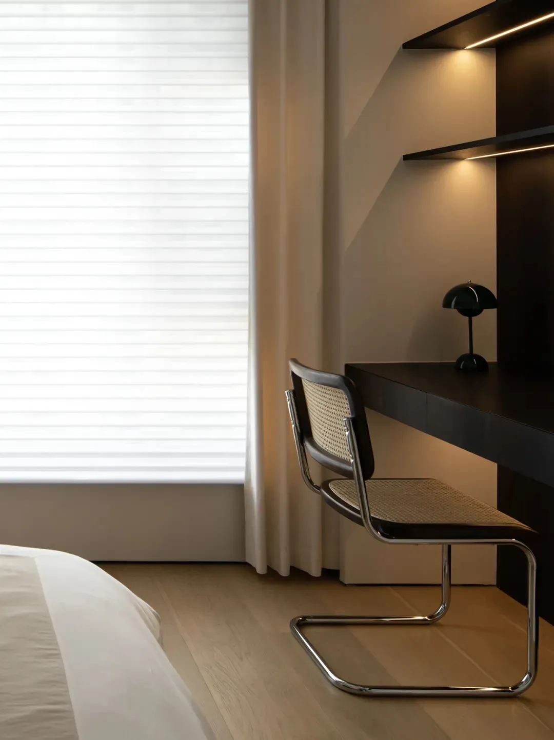 A minimalist workstation featuring a black desk and a woven chair with metal frame, placed by a window with blinds, against a