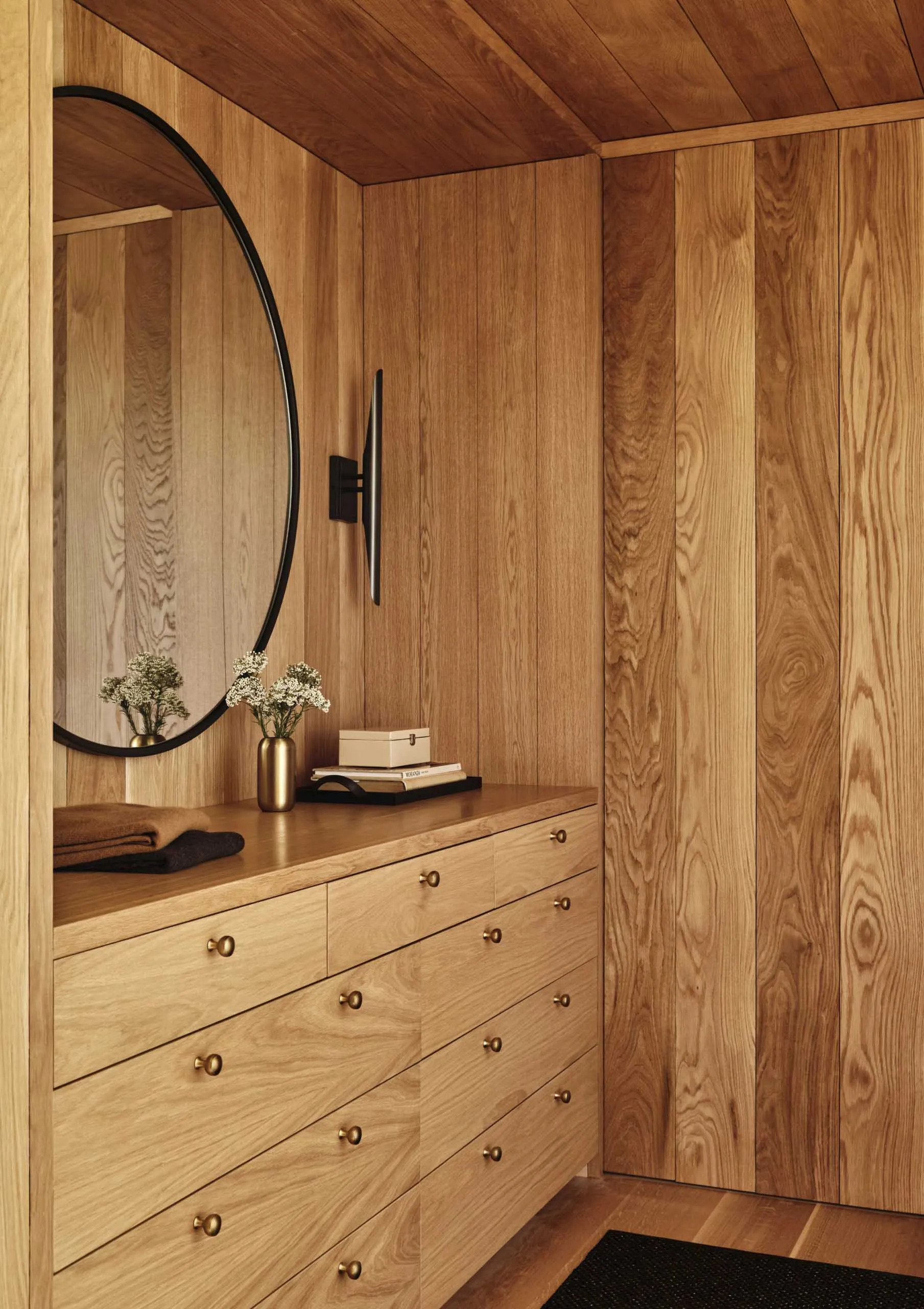 A minimalist wardrobe space featuring a wooden cabinet with multiple drawers and a circular mirror on a wooden wall.