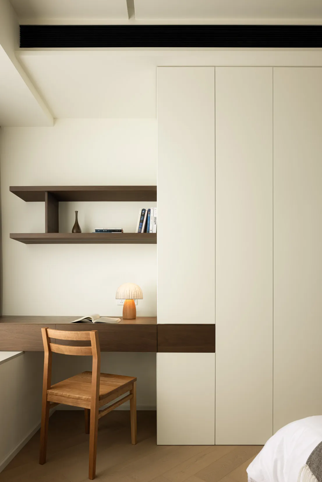 A minimalist bedroom with a wooden chair, white desk lamp, and white bookshelf.