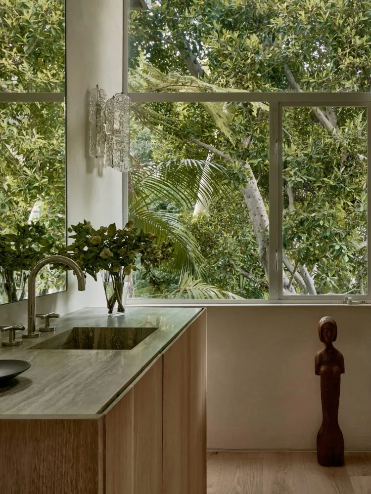 Modern residential bathroom featuring wooden furniture, marble countertop, and large windows overlooking lush greenery.