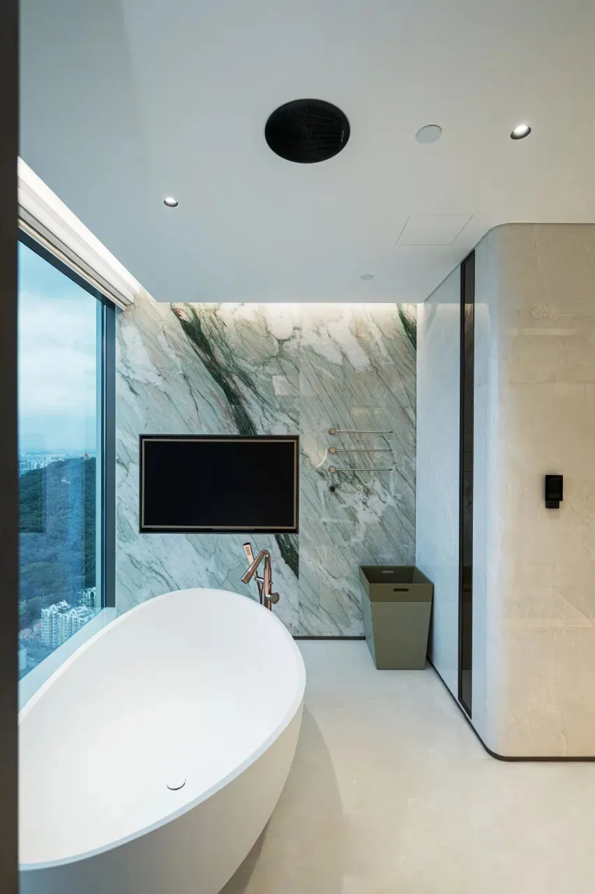 A modern residential bathroom featuring marble and concrete elements, including a freestanding bathtub, a television mounted