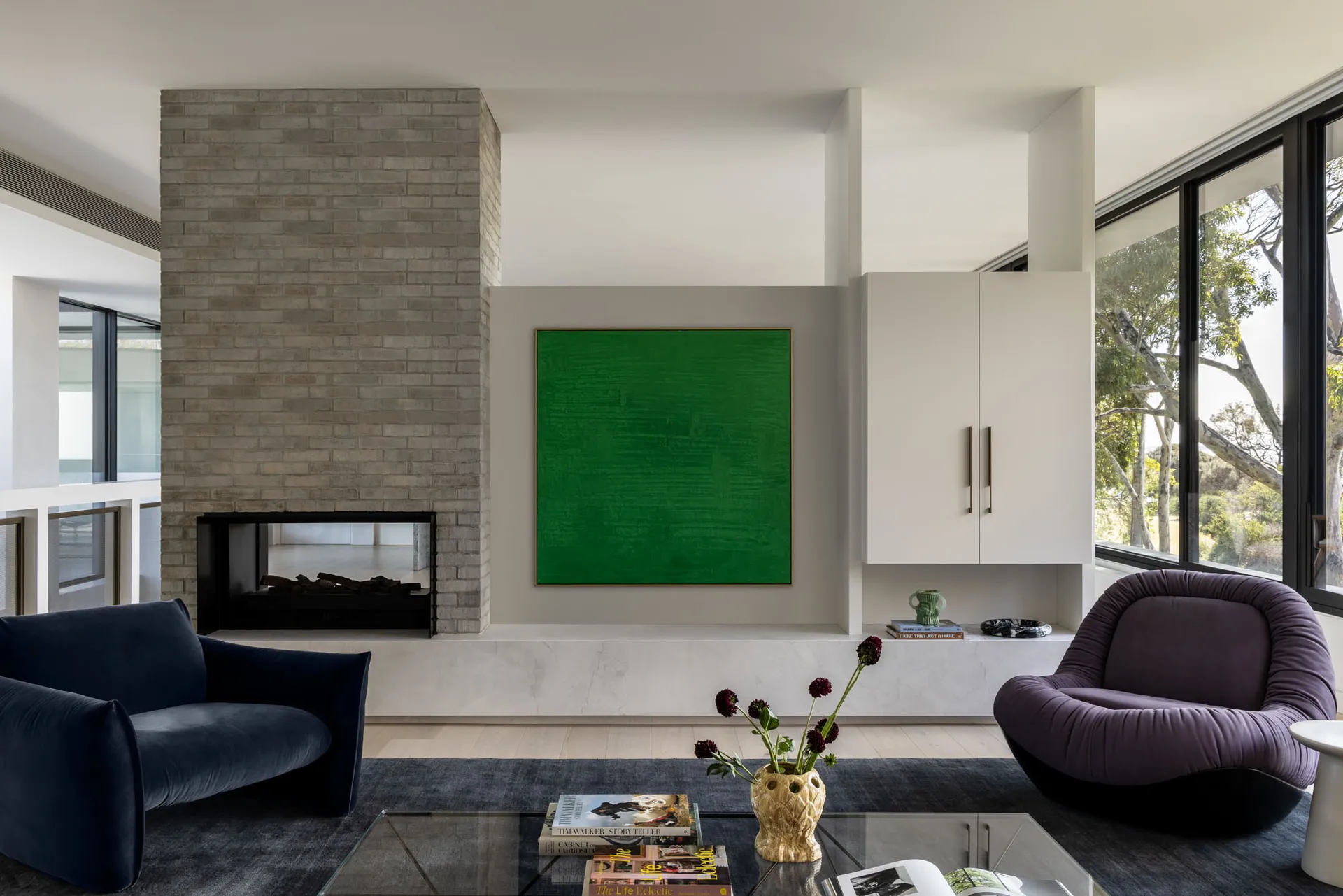 A modern contemporary living room featuring a green canvas art piece, dark blue and purple plush chairs, a glass coffee table
