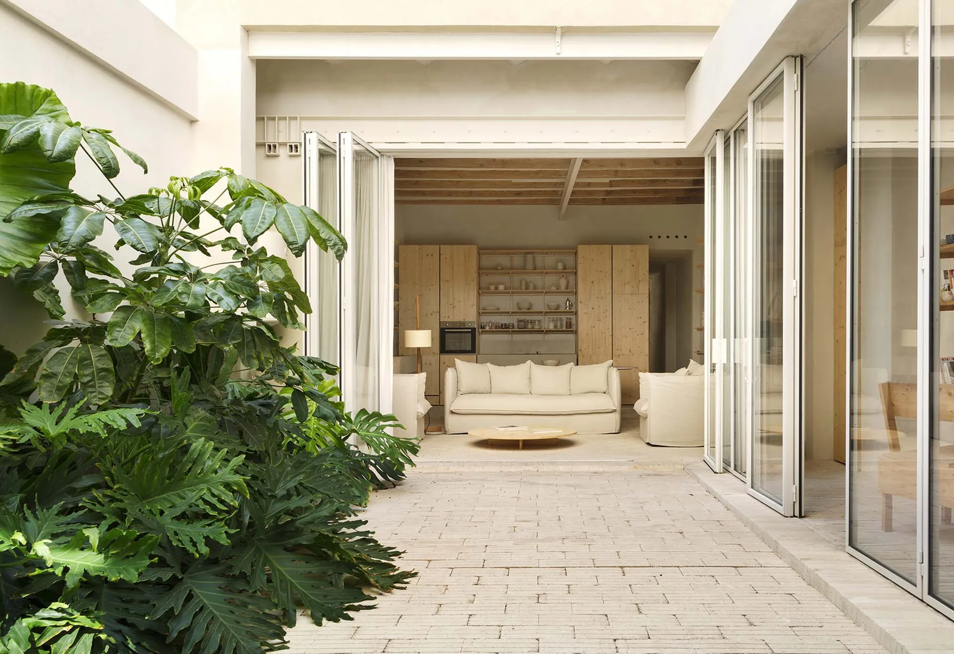 A minimalist living space with wooden furniture and open-plan design, featuring a white brick patio and large green plants.