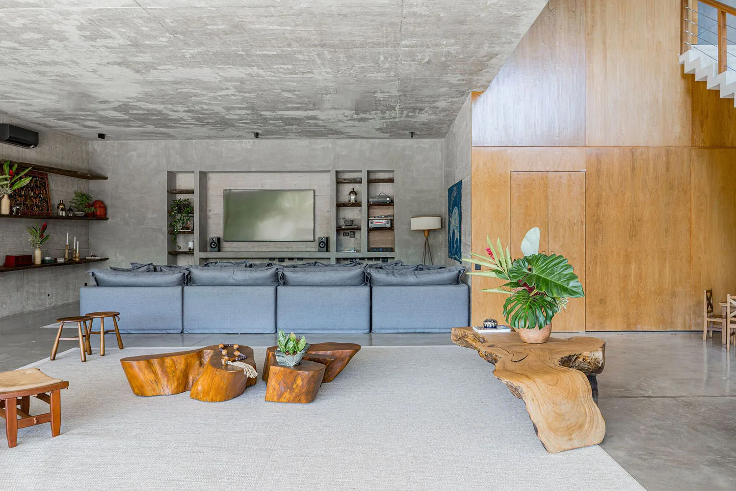An industrial living room with concrete floors and walls, featuring a large gray sectional sofa, wooden coffee tables, and a