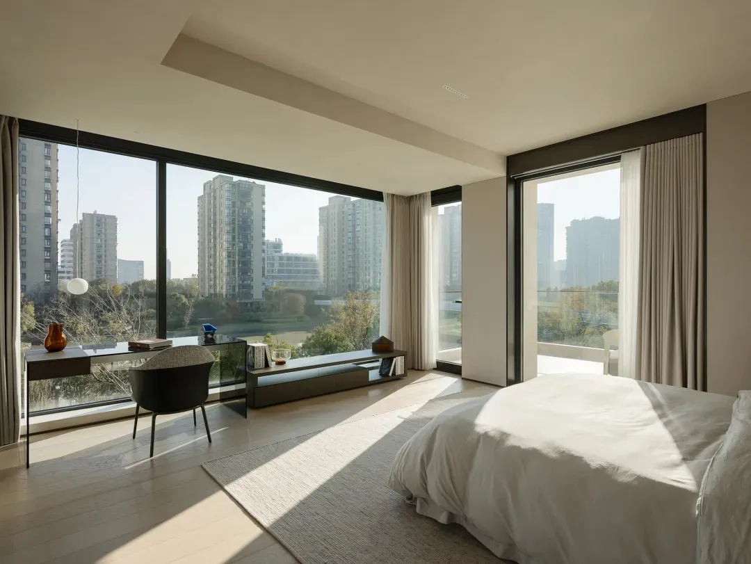 A minimalist bedroom with a large window overlooking city buildings and trees, featuring a bed with neutral linens and a simp