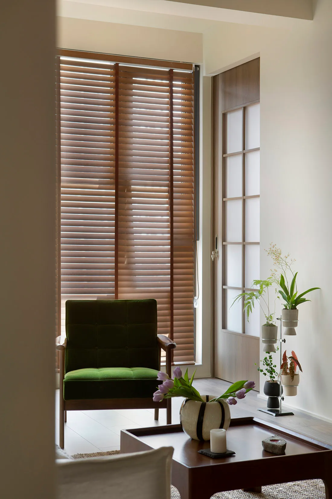 A minimalist living room with wooden blinds, a green accent chair, and a modern coffee table with plants and a vase of tulips