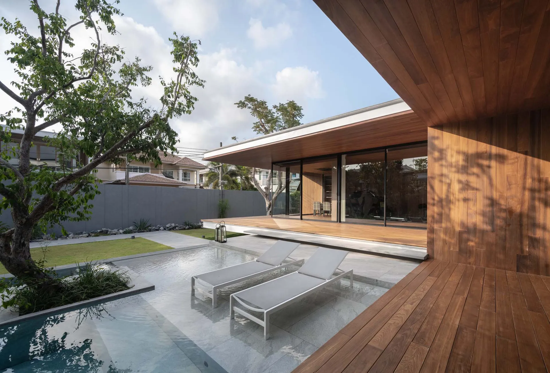 Modern residential home with wooden deck, glass walls, and concrete patio featuring outdoor furniture and a swimming pool sur