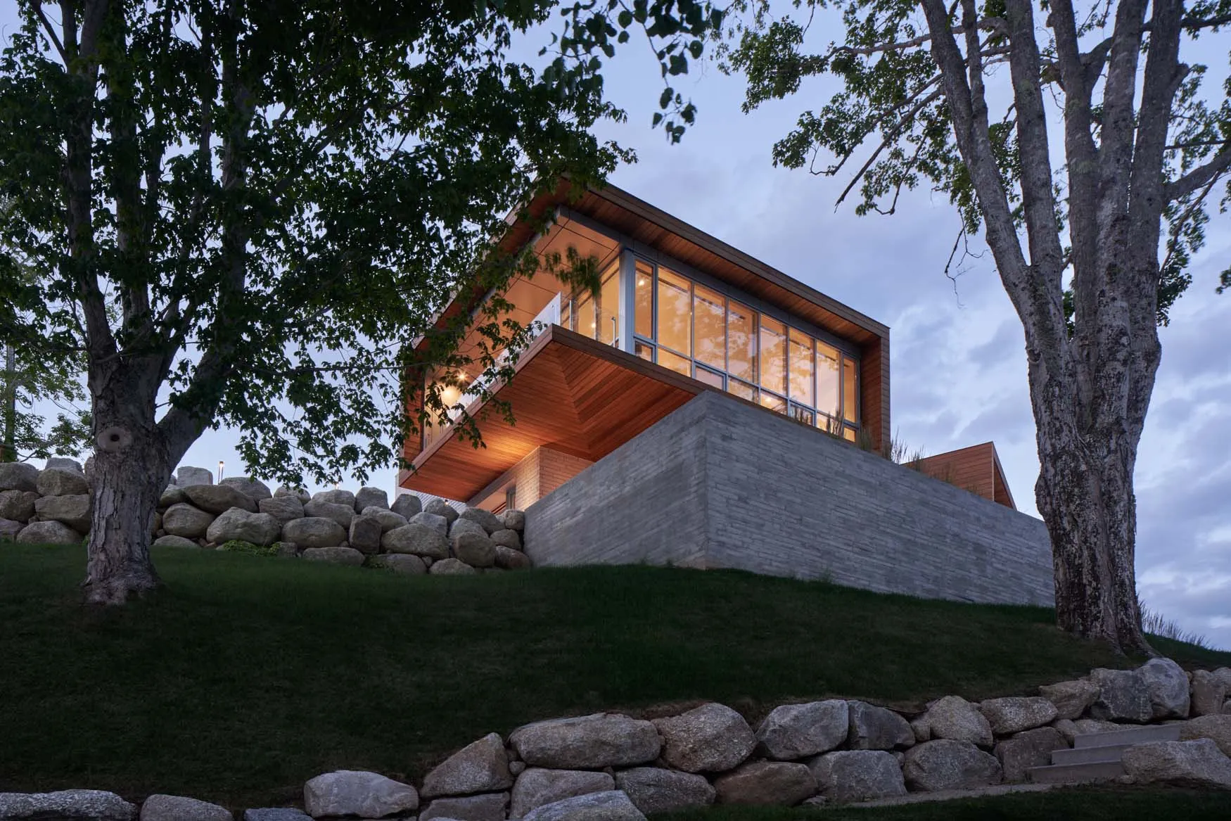 A modern house with wood exterior and concrete foundation, featuring large windows and a sloped roof.
