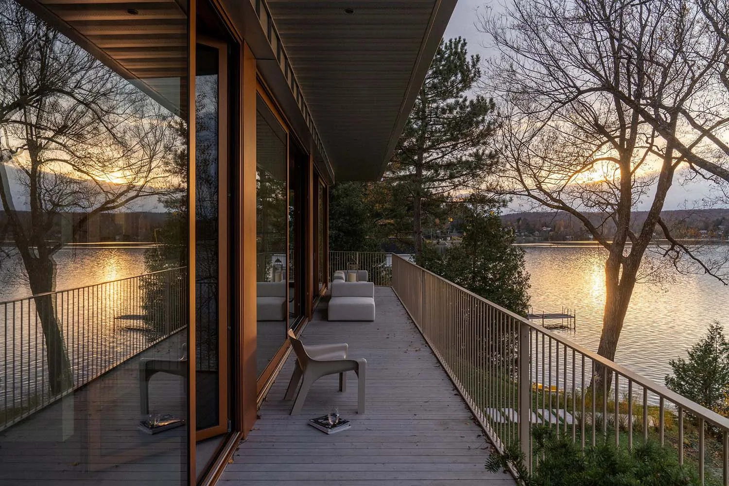 Modern exterior of a waterfront residence with wooden deck and furniture, overlooking a serene lake with sunset reflections.