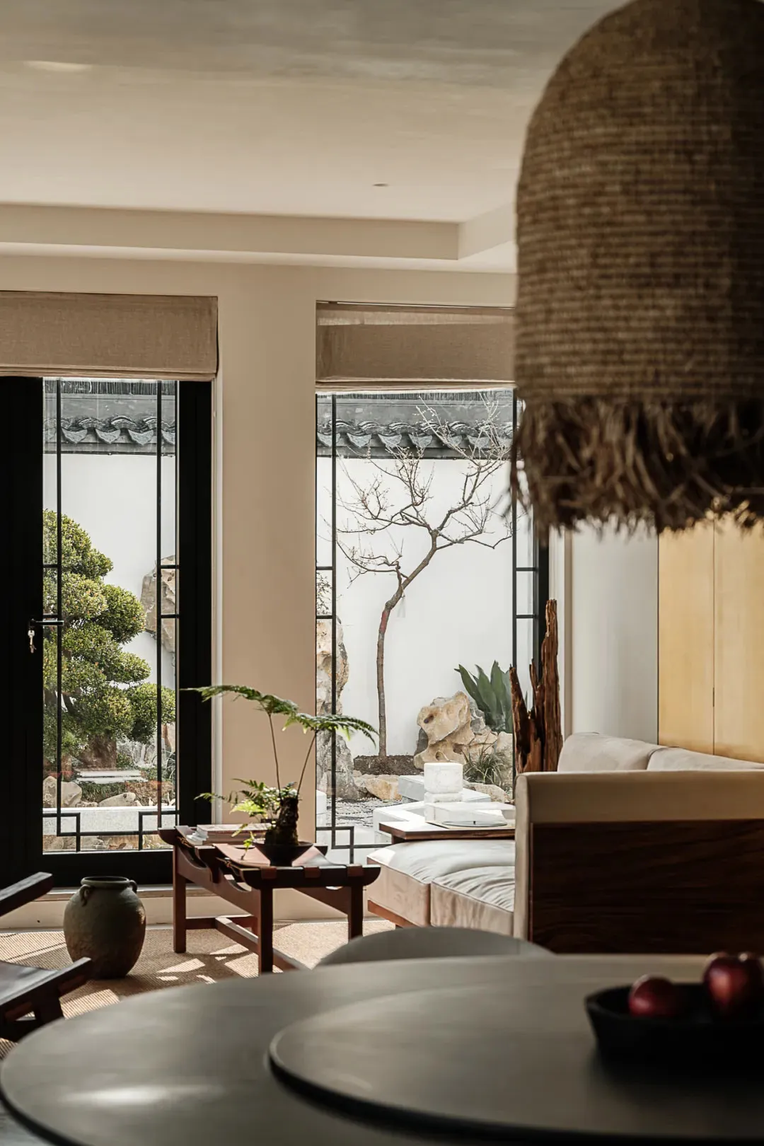 A minimalist living room with neutral-colored walls and furniture, featuring a large window with a view of a garden.