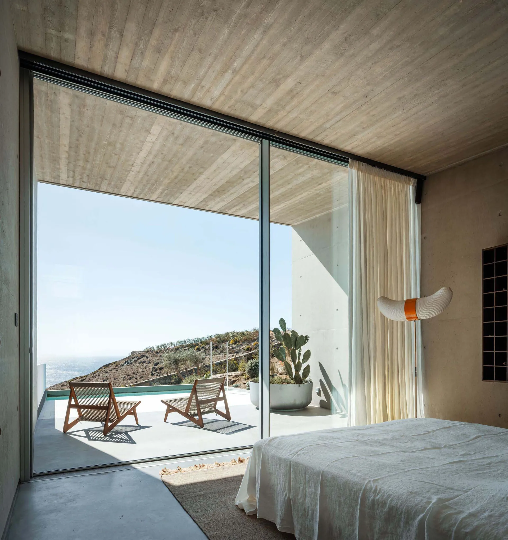 Modern minimalist bedroom featuring white furnishings, concrete walls, and a view of the ocean.