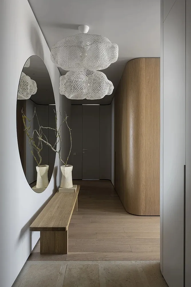 Modern minimalistic entryway with large wooden bench, circular mirrors, and glass chandelier with floral design.