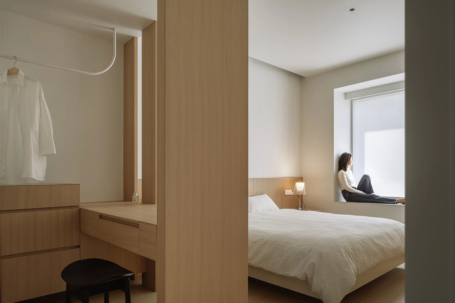 A minimalist bedroom featuring a white bed, a wooden desk, and a window with blinds.