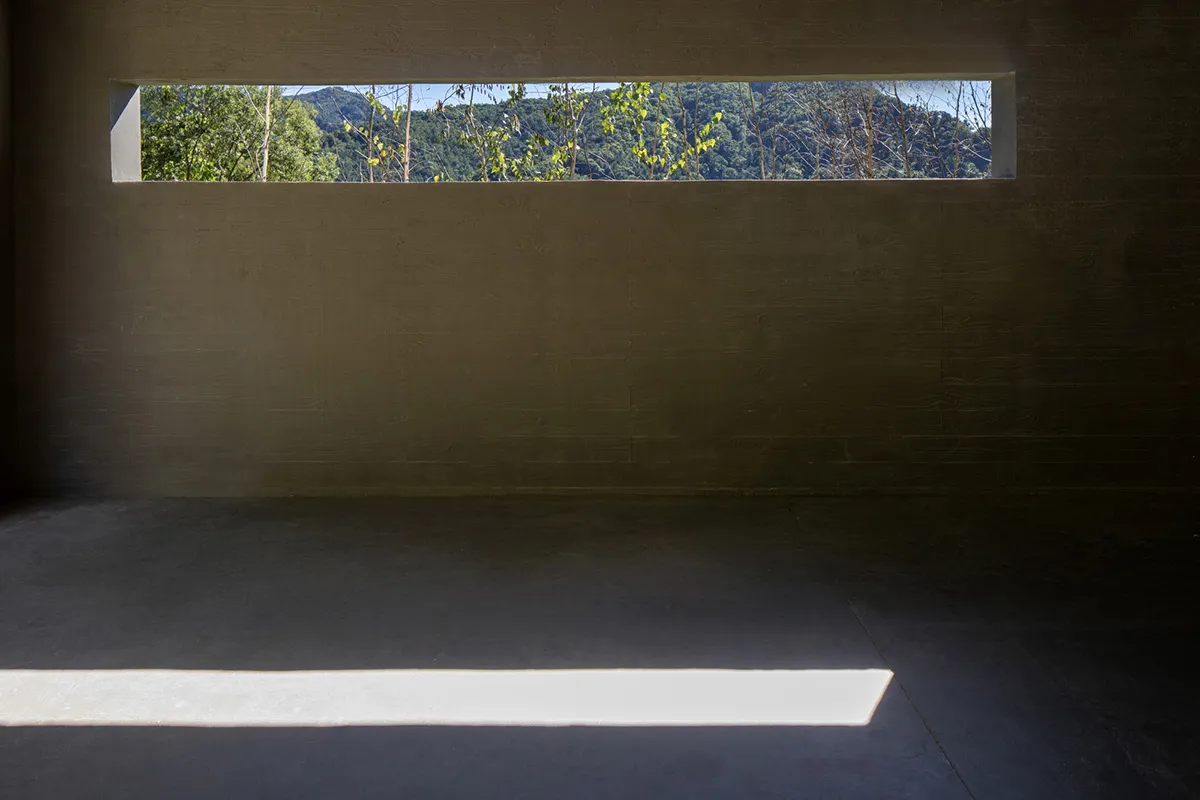 A minimalist interior space with a concrete floor and a small, rectangular window overlooking trees and mountains.