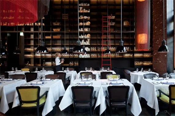 Modern restaurant interior with chairs and tables set with white cloth, multiple pendant lights, and red brick walls.