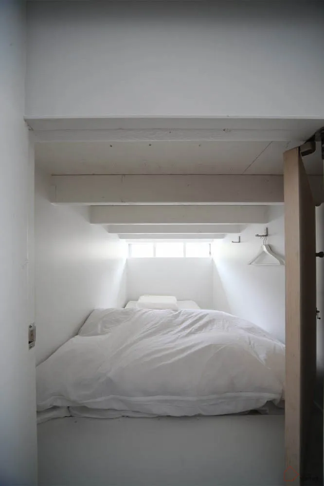 A minimalist bedroom with a bed covered in white textiles and a small window on the ceiling.