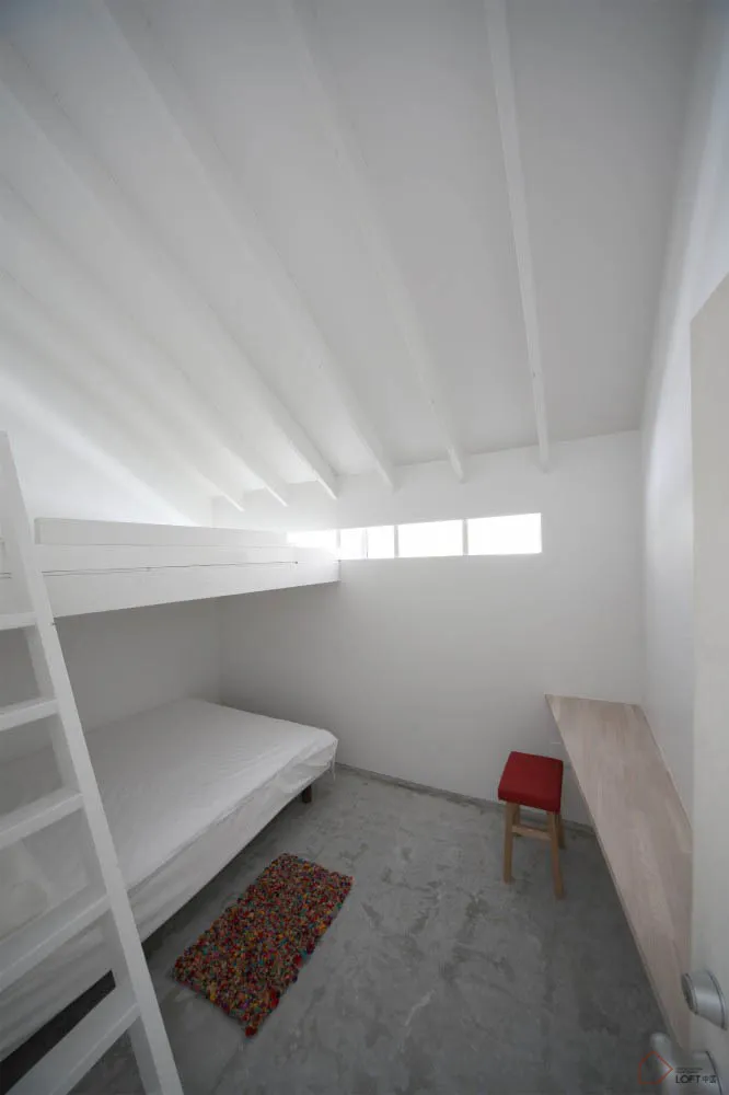 A minimalist bedroom features a concrete floor, bunk bed, and small desk with a red stool.